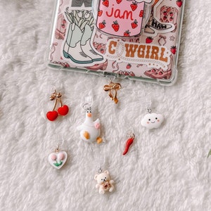 Puede incluir: Un conjunto de siete abalorios coloridos que incluyen un pato blanco, un chile rojo, una nube blanca con una cara sonriente, un corazón blanco con dos flores rosas, un oso de peluche blanco que sostiene un corazón rosa, una cereza roja con un tallo dorado y un lazo dorado.