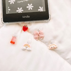 Könnte beinhalten: Nahaufnahme von fünf bunten Buch-Anhängern. Die Anhänger sind an einer durchsichtigen Plastikschnur befestigt und zeigen eine Erdbeere, eine rosafarbene Schleife, einen Regenbogen mit Wolken, einen rosafarbenen Gummibärchen und eine weiße Teigtasche mit einem lächelnden Gesicht. Die Anhänger hängen an einem schwarzen Kindle E-Reader mit einer durchsichtigen Hülle. Der Text auf dem Kindle lautet "support kindle" und "Property of Hays' Book Babes".