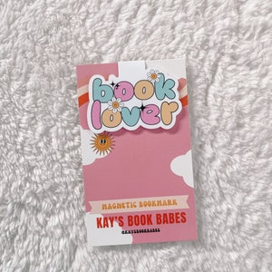 Może przedstawiać: Magnetyczny zakładka w kolorze r&oacute;żowym i białym z tekstem "book lover" w kolorowych literach w kształcie baniek. Zakładka zawiera r&oacute;wnież mały wz&oacute;r słońca i chmury. Na zakładce nadrukowano tekst "MAGNETIC BOOKMARK" i "KAY'S BOOK BABES". Na zakładce nadrukowano r&oacute;wnież nazwę profilu w mediach społecznościowych "@KAYSBooKBABES".