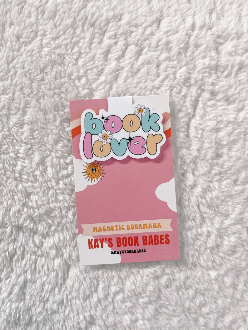 Może przedstawiać: Magnetyczny zakładka w kolorze r&oacute;żowym i białym z tekstem "book lover" w kolorowych literach w kształcie baniek. Zakładka zawiera r&oacute;wnież mały wz&oacute;r słońca i chmury. Na zakładce nadrukowano tekst "MAGNETIC BOOKMARK" i "KAY'S BOOK BABES". Na zakładce nadrukowano r&oacute;wnież nazwę profilu w mediach społecznościowych "@KAYSBooKBABES".