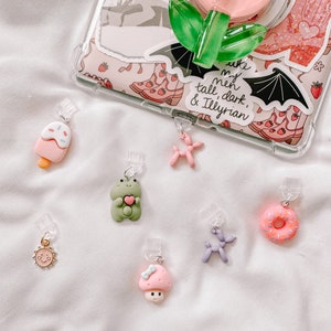 Puede incluir: Una colección de abalorios coloridos que incluyen un cono de helado rosa, una rana verde que sostiene un corazón, un sol amarillo, un hongo rosa con un lazo azul, un animal de globo morado, una rosquilla rosa con chispas y un animal de globo rosa.