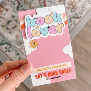 Może przedstawiać: R&oacute;żowo-biały magnes do zakładek z tekstem "book lover" w kolorowym, pękatym kroju pisma. Zakładka zawiera r&oacute;wnież słońce i chmury. Na zakładce nadrukowany jest tekst "KAY'S BOOK BABES @KAYSBOOKBABES".