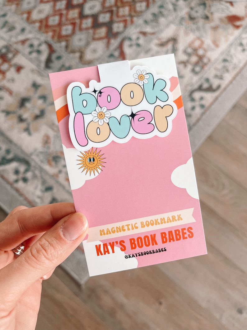 Może przedstawiać: R&oacute;żowo-biały magnes do zakładek z tekstem "book lover" w kolorowym, pękatym kroju pisma. Zakładka zawiera r&oacute;wnież słońce i chmury. Na zakładce nadrukowany jest tekst "KAY'S BOOK BABES @KAYSBOOKBABES".