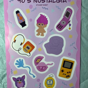 Könnte beinhalten: Ein Stickerbogen mit einem rosa Rand und dem Text "90's Nostalgia". Der Bogen zeigt Aufkleber einer Lavalampe, einer Troll-Puppe, eines Klapphandys, eines lila Stuhls, eines Furby, eines Tamagotchi, eines Game Boy, eines Kassettenrekorders und eines blauen, wellenförmigen Spielzeugs.