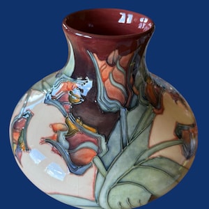 Puede incluir: Un jarrón de cerámica con cuerpo bulboso y borde acampanado. El jarrón presenta un diseño floral en tonos naranja, rojo, verde y azul sobre un fondo crema. El borde es de color burdeos oscuro.