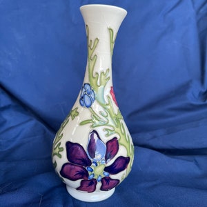 Pode incluir: Um vaso de cerâmica branco com um design floral. O vaso apresenta uma grande flor roxa, botões azuis e vermelhos e folhas verdes. O vaso tem um pescoço estreito e uma base larga.