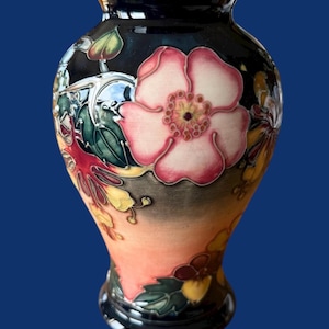 Puede incluir: Un jarrón de cerámica decorativo con base y borde azul oscuro, con un gran diseño floral rosa y blanco. El jarrón tiene un degradado de tonos naranja y amarillo, con hojas verdes y detalles florales. El jarrón mide unos 20 cm de alto.