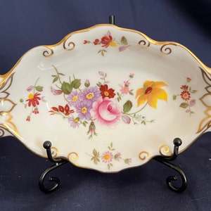 Puede incluir: Un plato ovalado blanco con un borde dorado y un diseño floral. El plato tiene un delicado patrón floral en tonos de rosa, amarillo, rojo y morado. El plato está sobre un soporte de metal negro.