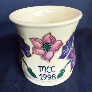 Puede incluir: Una taza de cerámica blanca con un esmalte craquelado presenta tres flores pintadas a mano en tonos rosa, morado y verde. La taza tiene el texto "MCC 1998" en la parte inferior.