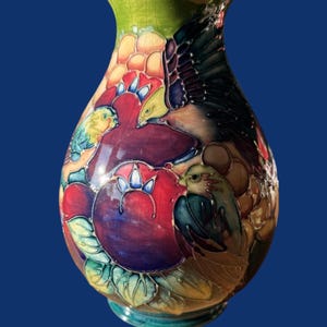 Puede incluir: Un jarrón de cerámica con forma bulbosa y acabado brillante con textura agrietada. La superficie está decorada con un colorido diseño en relieve de pájaros, grandes frutas rojas y moradas, y hojas verdes sobre un fondo vibrante y multicolor.