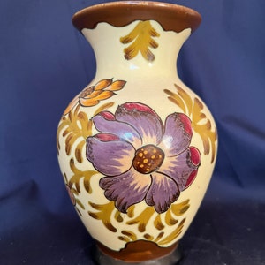 Puede incluir: Un jarrón de cerámica de color crema con un borde y una base marrones. El jarrón está decorado con un patrón floral en tonos de morado, amarillo, naranja y rojo.