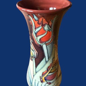 Puede incluir: Un jarrón decorativo alto con borde acampanado y base bulbosa. El jarrón es principalmente burdeos con un diseño floral en tonos naranja, azul y verde. El diseño parece estar pintado a mano o esmaltado sobre la superficie cerámica.
