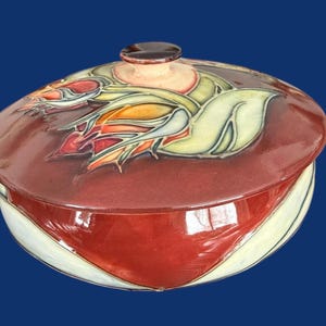 May include: A ceramic lidded serving dish with a maroon base and lid. The lid features a floral design in shades of red, orange, and yellow, with a small brown knob. The dish has a white and cream-coloured accent on the sides.