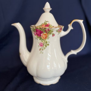 Könnte beinhalten: Weiße Porzellan-Teekanne mit goldener Verzierung und einem Blumenmuster aus rosa, gelben und roten Rosen. Die Teekanne hat einen Deckel mit einem Knopf oben.