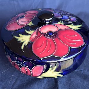 Könnte beinhalten: Eine blaue und weiße Keramik-Schmuckdose mit Deckel. Die Dose ist mit roten und rosa Blumen und grünen Blättern verziert.