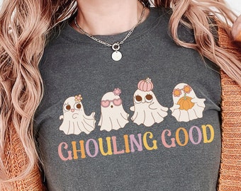 Könnte beinhalten: Dunkelgraues T-Shirt mit einem skurrilen Design von vier Cartoon-Geistern mit Accessoires und dem Text "Ghouling Good" in bunten Buchstaben. Die Geister sind mit Blumen, Sonnenbrillen und Kürbissen geschmückt. Über dem Shirt wird eine orangefarbene Strickjacke getragen.