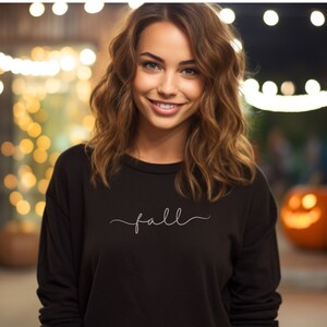 Puede incluir: Una mujer con una sudadera negra de manga larga con la palabra "fall" en cursiva. El fondo presenta luces borrosas y calabazas talladas, sugiriendo un tema de otoño o Halloween.
