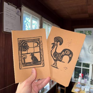 Peut inclure: Deux petits carnets marrons avec des illustrations à l'encre noire. Un carnet représente un chat regardant par une fenêtre avec une bougie et une plante. L'autre carnet représente un coq avec un motif soleil et lune.