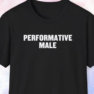May include: A black short-sleeved t-shirt featuring the text PERFORMATIVE MALE printed in white capital letters across the chest. The garment has a crew neckline and a standard fit, showcasing a simple design with bold, centered typography.