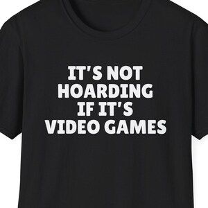 Può includere: T-shirt nero con la scritta bianca "IT'S NOT HOARDING IF IT'S VIDEO GAMES". La maglietta a maniche corte ha lo scollo tondo. Il testo è centrato sulla parte anteriore della maglietta.