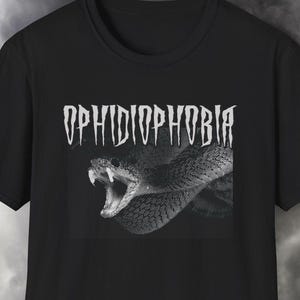 Könnte beinhalten: Schwarzes T-Shirt mit dem Wort "OPHIDIOPHOBIA" in Weiß, über einem Graustufenbild einer Schlange mit offenem Maul, Zähne sichtbar. Der Hintergrund ist wolkig, dunkelgrau.
