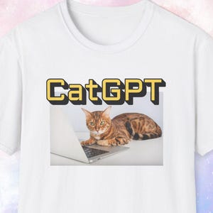 Könnte beinhalten: Weißes T-Shirt mit einer Grafik einer Katze, die einen Laptop benutzt, und dem Text "CatGPT". Die Katze hat braune und schwarze Markierungen. Der Text ist in Gelb und Schwarz. Das T-Shirt ist aus weichem Material.