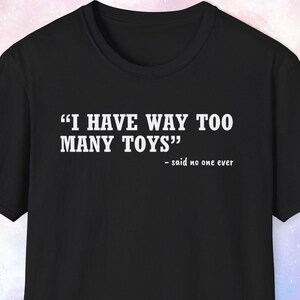 May include: A black short-sleeved t-shirt featuring white text printed on the chest that reads "I HAVE WAY TOO MANY TOYS" followed by the smaller text "- said no one ever" in a clean, sans-serif font style against the solid black fabric.