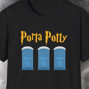 Puede incluir: Camiseta negra con la frase "Porta Potty" en letras doradas sobre un gráfico de tres baños portátiles azules. El diseño está centrado en la parte delantera de la camiseta.