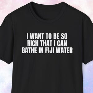 May include: Black t-shirt with white text that reads: "I WANT TO BE SO RICH THAT I CAN BATHE IN FIJI WATER". The shirt has short sleeves and a crew neck.
