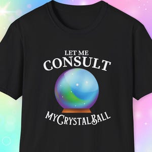 May include: Black t-shirt with the text "LET ME CONSULT MY CRYSTAL BALL" in white. The design features a colorful crystal ball on a wooden stand. The shirt is a classic crew neck style.