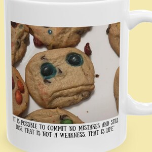 Può includere: Una tazza in ceramica bianca con una stampa raffigurante diversi biscotti con gocce di cioccolato, uno dei quali ha un volto con occhi blu. Sotto l'immagine, il testo recita: IT IS POSSIBLE TO COMMIT NO MISTAKES AND STILL LOSE. THAT IS NOT A WEAKNESS. THAT IS LIFE.