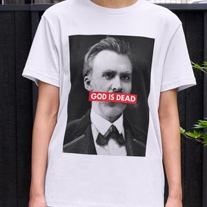 May include: A white t-shirt featuring a black and white portrait of a man with a bow tie and the text "GOD IS DEAD" in red. The person is wearing a black skirt and standing outside.