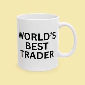 May include: White ceramic mug with the text "WORLD'S BEST TRADER" in bold black letters. The mug has a curved handle and is set against a light yellow background. A simple, yet bold design.
