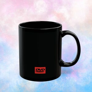 May include: A black ceramic mug with a curved handle. The mug features the red "DVD VIDEO" logo on the side. The background is a soft blend of pink, blue, and white.