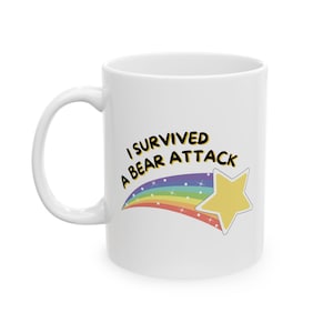 Puede incluir: Taza de cerámica blanca con el texto "I Survived a Bear Attack" en negro y amarillo. Un arcoíris y una estrella amarilla también están en la taza. La taza tiene un asa blanca.