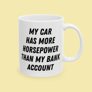May include: A white ceramic mug with a handle, featuring the black printed text MY CAR HAS MORE HORSEPOWER THAN MY BANK ACCOUNT. The mug has a cylindrical shape and a smooth finish, displaying the bold sans-serif lettering clearly on the front.