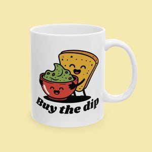 May include: White ceramic mug featuring a cartoon illustration of a bowl of guacamole and a piece of toast, both with smiling faces. The text "Buy the dip" is printed in black. The mug has a curved handle.