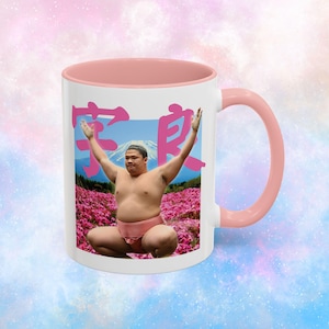 May include: White ceramic mug with a pink interior and handle. The mug features a sumo wrestler graphic with Japanese text, set against a backdrop of Mount Fuji and pink flowers. The design wraps around the mug.