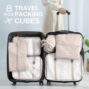 May include: Open suitcase filled with eight beige travel packing cubes. The cubes are various sizes and shapes, including zippered bags and pouches. The text "8 TRAVEL PCS PACKING CUBES" is displayed above the suitcase.