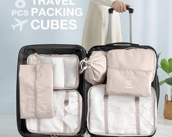 May include: Open suitcase filled with eight beige travel packing cubes. The cubes are various sizes and shapes, including zippered bags and pouches. The text "8 TRAVEL PCS PACKING CUBES" is displayed above the suitcase.