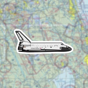 May include: A sticker depicting the NASA Space Shuttle Columbia in white and black. The shuttle has the words "NASA" and "United States" printed on it. The sticker is set against a map background.