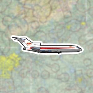 Pode incluir: Um autocolante com um design de avião TWA vintage. A aeronave é principalmente cinzenta com detalhes em vermelho e branco, e o logótipo da TWA é exibido na cauda e na fuselagem. O autocolante tem uma borda branca.