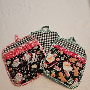 May include: Three Christmas-themed quilted pot holders. Each pot holder features a black background with Santa Claus, gingerbread men, and candy cane designs. The edges are trimmed with red, green, and black and white checkered fabric, and each has a loop for hanging.