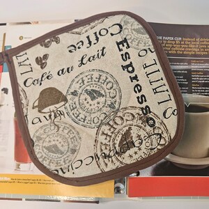 May include: A brown and beige pot holder with a coffee theme. It has the words "Cafe au Lait", "Espresso", and "Coffee" printed on it. The pot holder is on top of a cookbook with a cup of coffee and sugar cubes.