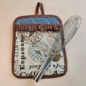 May include: Brown quilted pot holder with a blue and white checkered pattern and a brown fringe. The pot holder has the words "Espresso", "Coffee", and "Café Lait" printed on it. A silver whisk is lying on top of the pot holder.