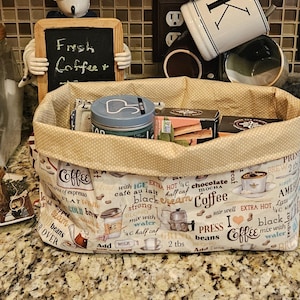 May include: A rectangular fabric basket with a coffee-themed print, filled with coffee and tea supplies. The basket has a beige polka-dot lining and features text like "Coffee" and "Fresh Coffee+".