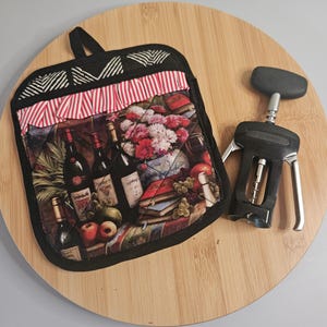 May include: A black quilted hot pad with a red and white striped trim features a colorful image of wine bottles, grapes, and flowers. A black and silver corkscrew is also pictured.