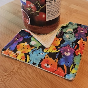 May include: A colorful cat-themed coaster with a black background. The coaster features a repeating pattern of cartoon cats in various colors, including orange, blue, and purple. The coaster is made of fabric and has a white border.