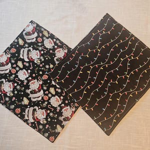 May include: Two square Christmas-themed fabric napkins. One features a black background with Santa Claus, candy canes, and cookies. The other has a black background with strings of colorful Christmas lights.