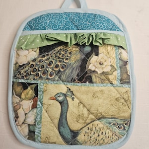 May include: A quilted pot holder featuring a peacock design. The pot holder has a light blue border and a hanging loop. The main design includes images of peacocks, flowers, and a green ruffled trim. The pot holder is approximately 8 inches wide and 9 inches tall.